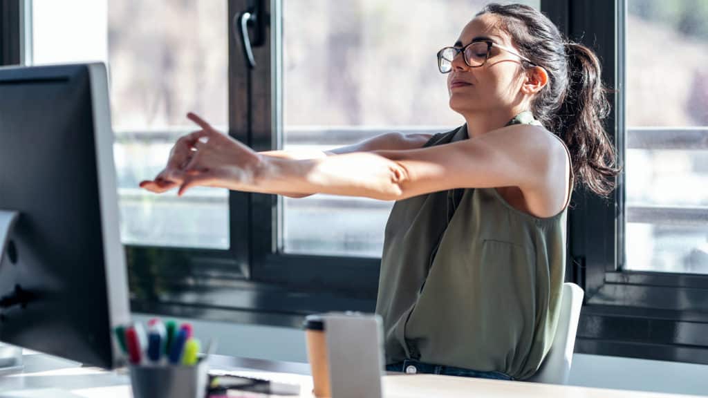 imagem de mulher branca com cabelo presos, óculos, sentada em uma mesa em frente a um computador enquanto faz alongamento para home office.